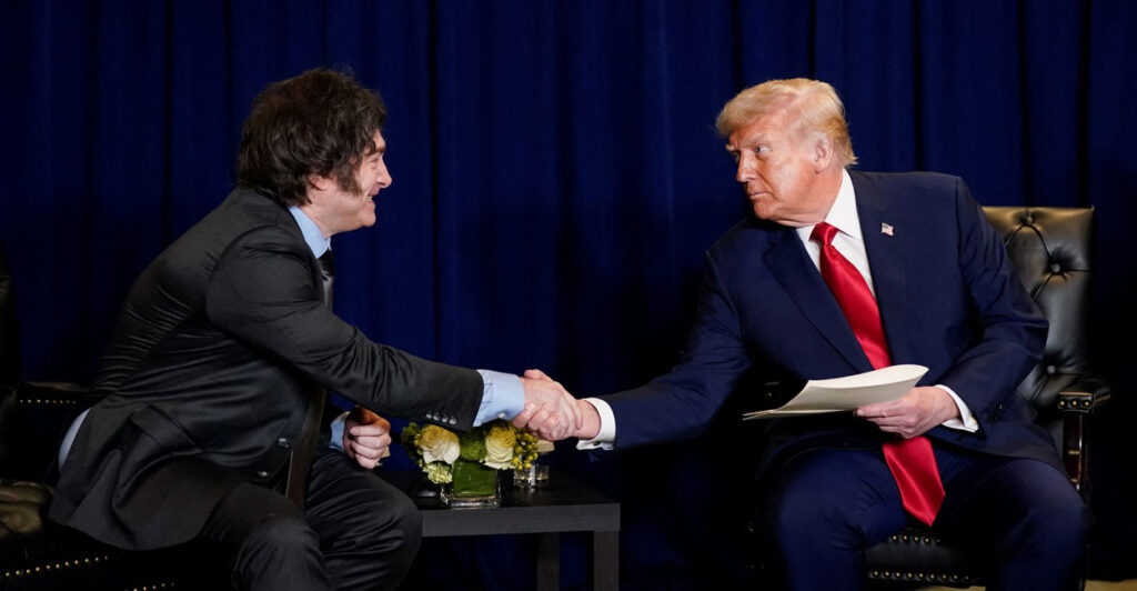 President Donald Trump shakes hands with Argentina's President Javier Milei as they sit in front of a blue curtain.