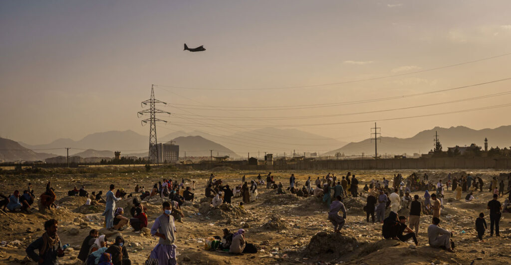 A military transport flies over an Afghan field, as Afghans watch below.