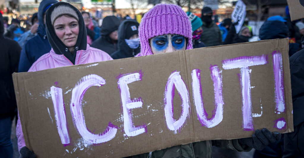 A protester with purple knit hat and blue makeup around eyes holds sign reading "ICE OUT"