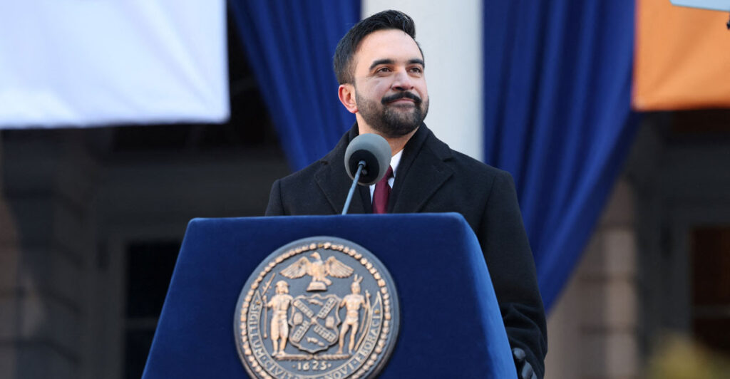 Zohran Mamdani behind a blue podium.