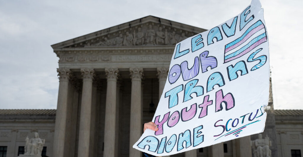 A sign held up reading "Leave Our Trans Youth Alone" against backdrop of Supreme Court.