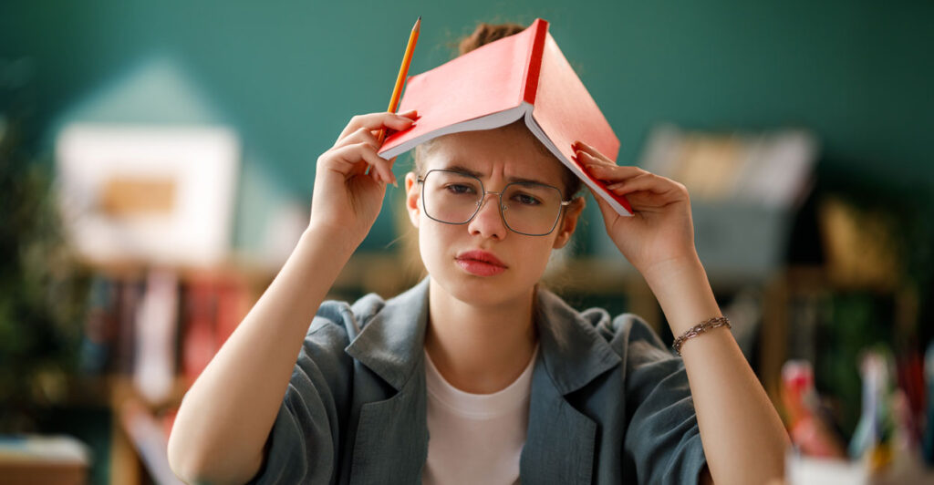 Young befuddled girl with glasses holds open book on top of her head in classroom.