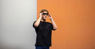 Mark Zuckerberg, in a black t-shirt, puts on a pair of dark glasses against a half-white, half-orange background.
