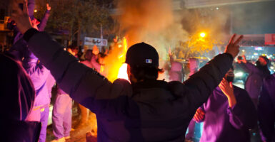 Iranian protester with back to the camera, arms outstretched, giving peace signs, amid nighttime protest. Flames in front of him.