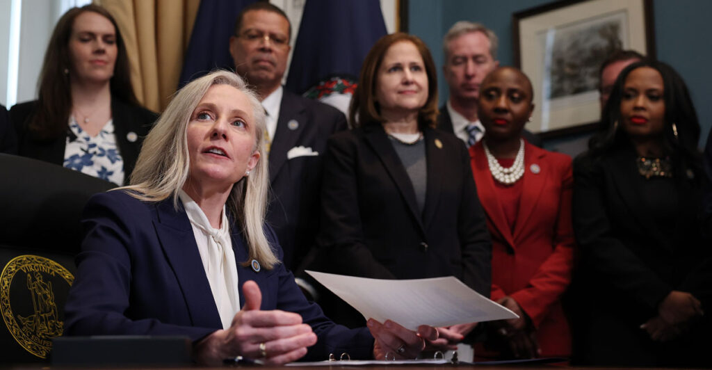 Virginia Gov. Abigail Spanberger at a desk, holding a document, as several people look on.