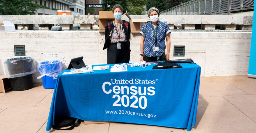 Two female masked 2020 Census workers stand behind a table outside Lincoln Center