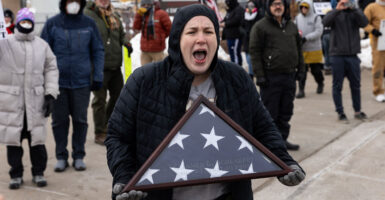 Woman in black winter coat with hoodie screams at ICE agents while holding folded American flag.