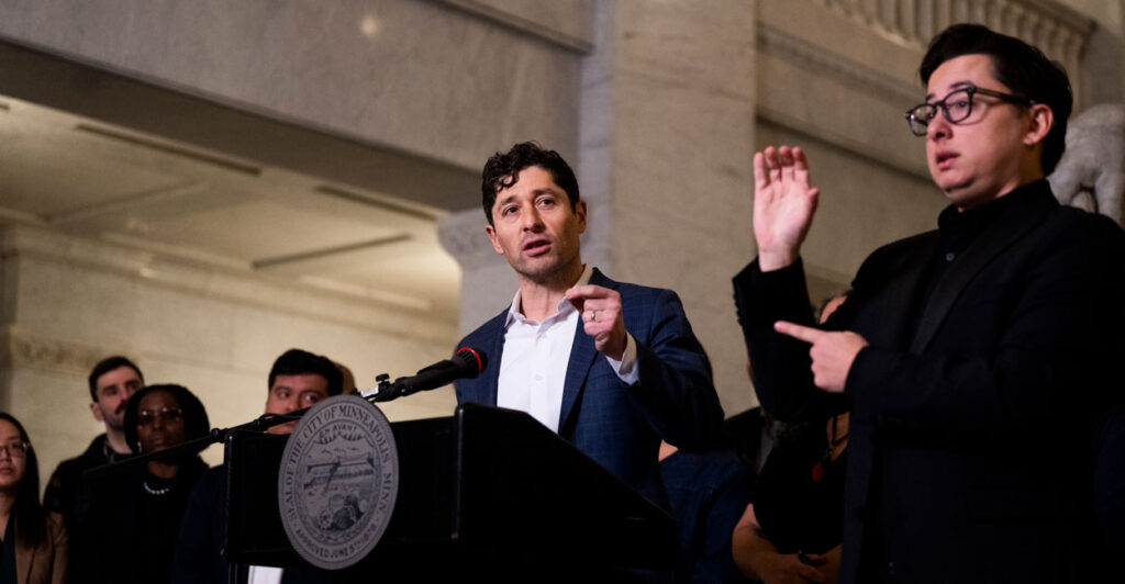 Minneapolis Mayor Jaco Frey speaks at a podium as an sign-language interpreter raises one hand and uses his left hand to point at Frey.
