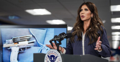 DHS Secretary Kristi Noem in a blue jacket speaks behind a podium next to a photo of a semi-automatic handgun.