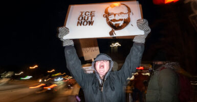 Woman in gray winter clothing screams as she holds up sign with image of Alex Pretti with caption ICE OUT NOW.