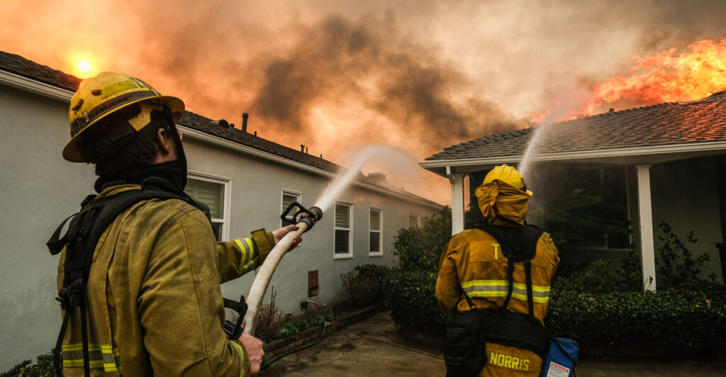 Firefighters coordinate their efforts, aiming hoses at a house with flames consuming its roof in Pacific Palisades, California, on January 8, 2025.