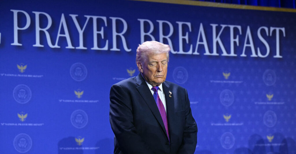 President Trump bows his head in prayer in front of blue "Prayer Breakfast" background.