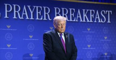 President Trump bows his head in prayer in front of blue "Prayer Breakfast" background.
