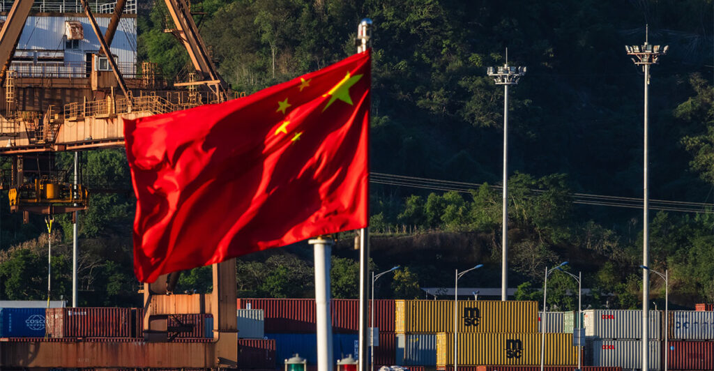 CHONGQING, CHINA - AUGUST 12: The national flag of China waves in the wind at a container terminal on the Yangtze River, with dockside cranes and stacked shipping containers visible in the background, on August 12, 2025 in Chongqing, China. (Photo by Cheng Xin/Getty Images)