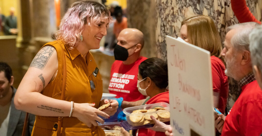 Transgender Minnesota state Rep. Leigh Finke, in brown shirt and pink died hair, greets kids in red shirts and masks.
