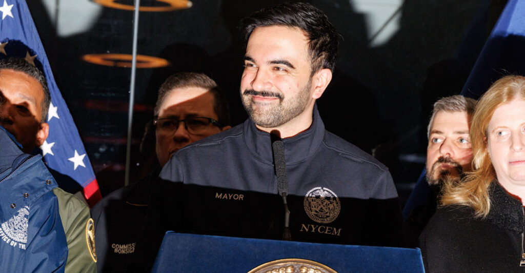 A smiling Zohran Mamdani in dark winter jacket behind a podium.