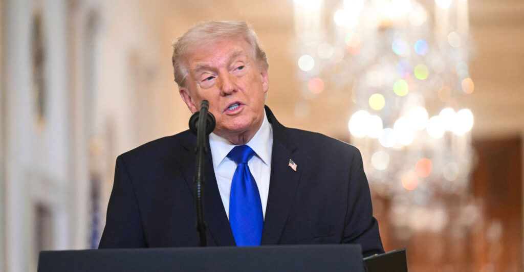 President Trump in dark suit and blue tie speaks at a podium in East Room of the White House.