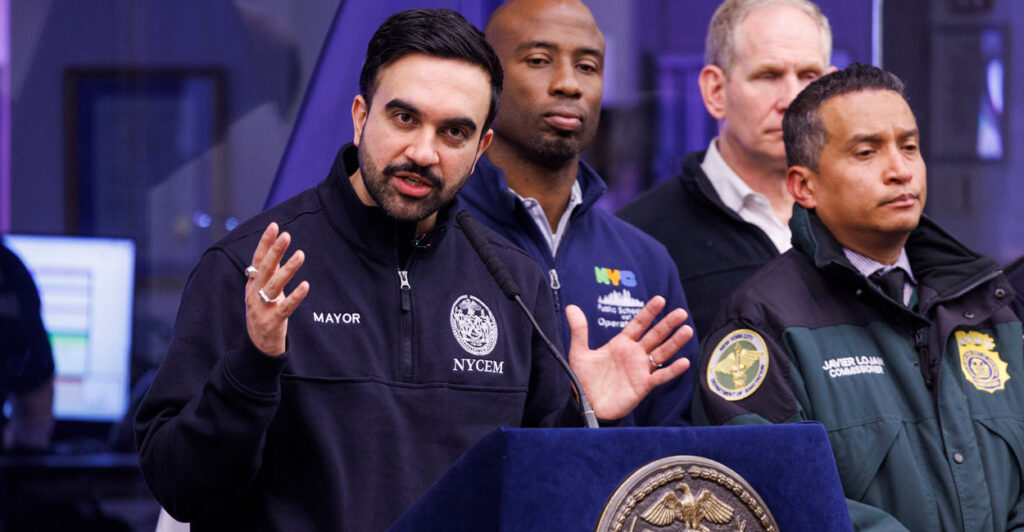 Zohran Mamdani in blue jacket stands at podium with arms outstretches and officials look on.