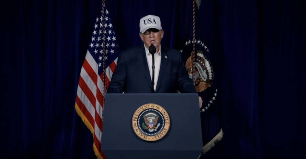 President Donald Trump behind a podium, in blue jacket, white shirt and "USA" cap, speaks.