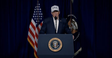 President Donald Trump behind a podium, in blue jacket, white shirt and "USA" cap, speaks.