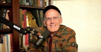 A smiling shot of Victor Davis Hanson seated at a microphone in a rust-colored flannel shirt.