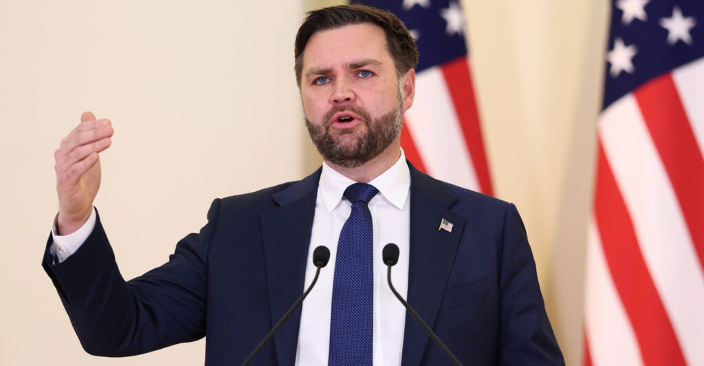 Vice President JD Vance gestures while he stands at a podium with American flags in the background.