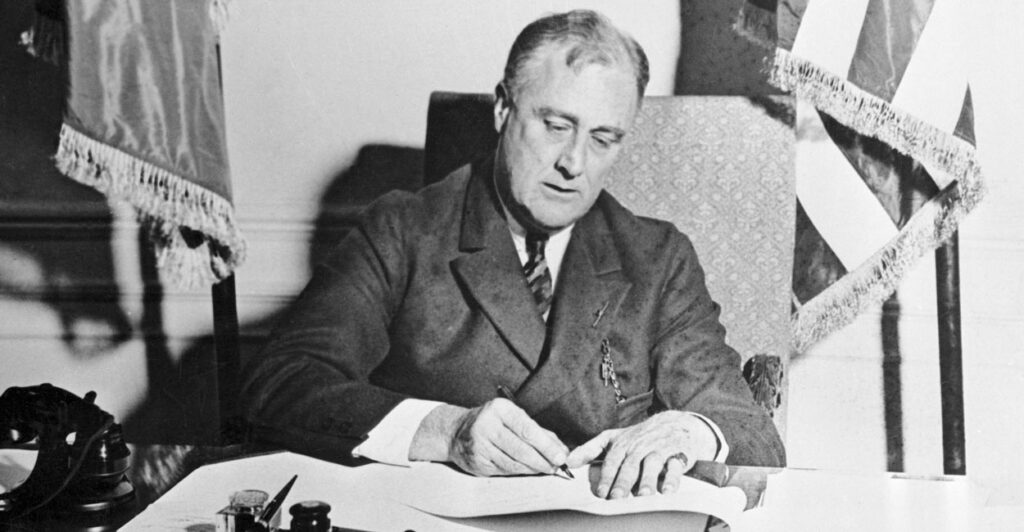 Black-and-white photo of President Franklin D. Roosevelt at desk signing legislation.