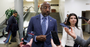 Sen. Raphael Warnock, in blue suit talks to reporters inside the U.S. Capitol.
