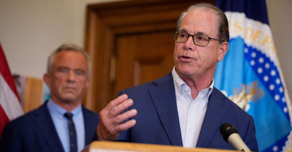 Indiana Gov. Mike Braun gestures while speaking at a podium, HHS Secretary Robert F. Kennedy Jr. behind him.