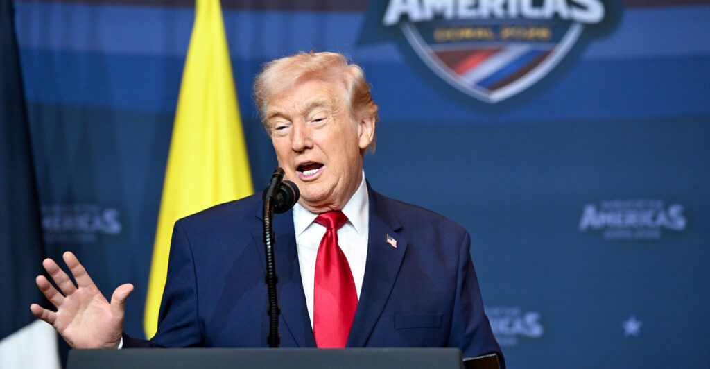President Trump gestures with his right hand while speaking at a podium in front of a "Shield of America" sign.
