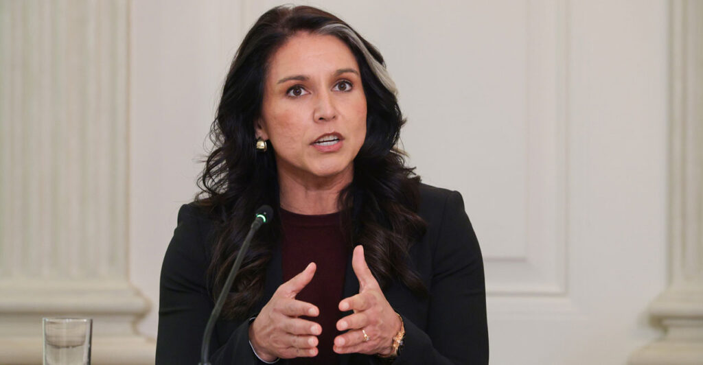Tulsi Gabbard, in black, brings hands together as she speaks against a white background.