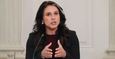 Tulsi Gabbard, in black, brings hands together as she speaks against a white background.