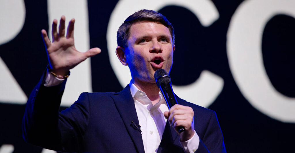 James Talarico in dark jacket and open white shirt raises right hand while delivering a speech.