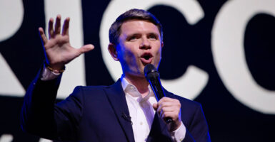 James Talarico in dark jacket and open white shirt raises right hand while delivering a speech.
