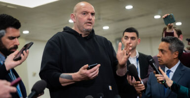 John Fetterman in black hoodie holds cellphone while talking to reporters.
