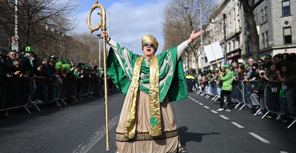 An actor depicting St. Patrick spreads his arms wide in the middle of a Dublin street as parade spectators look on.