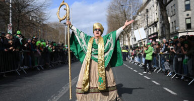 An actor depicting St. Patrick spreads his arms wide in the middle of a Dublin street as parade spectators look on.