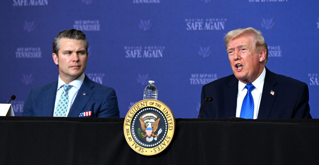 Secretary of War Pete Hegseth watches President Donald Trump speak in front of a blue backdrop with the words in white "Make America Safe Again."