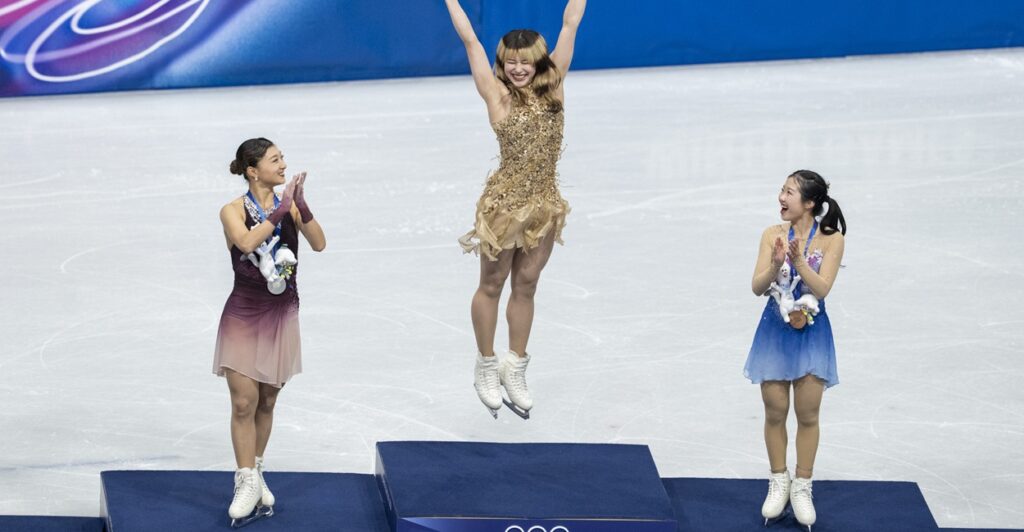 Alysa Liu leaping in the air on the Olympic podium as two other skaters applaud.