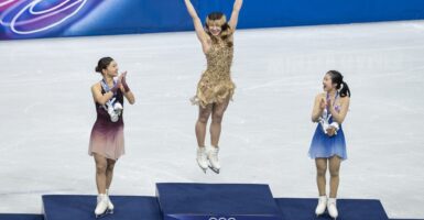 Alysa Liu leaping in the air on the Olympic podium as two other skaters applaud.