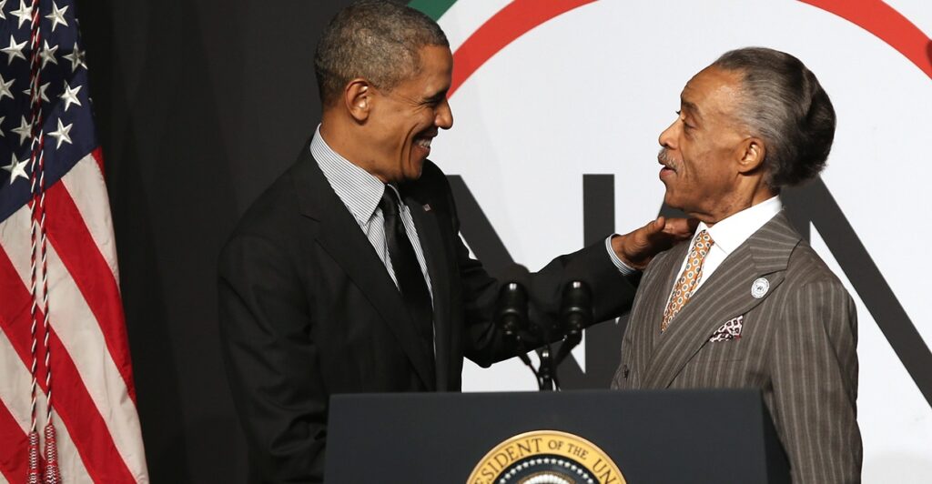 President Barack Obama greets Rev. Al Sharpton on a podium.