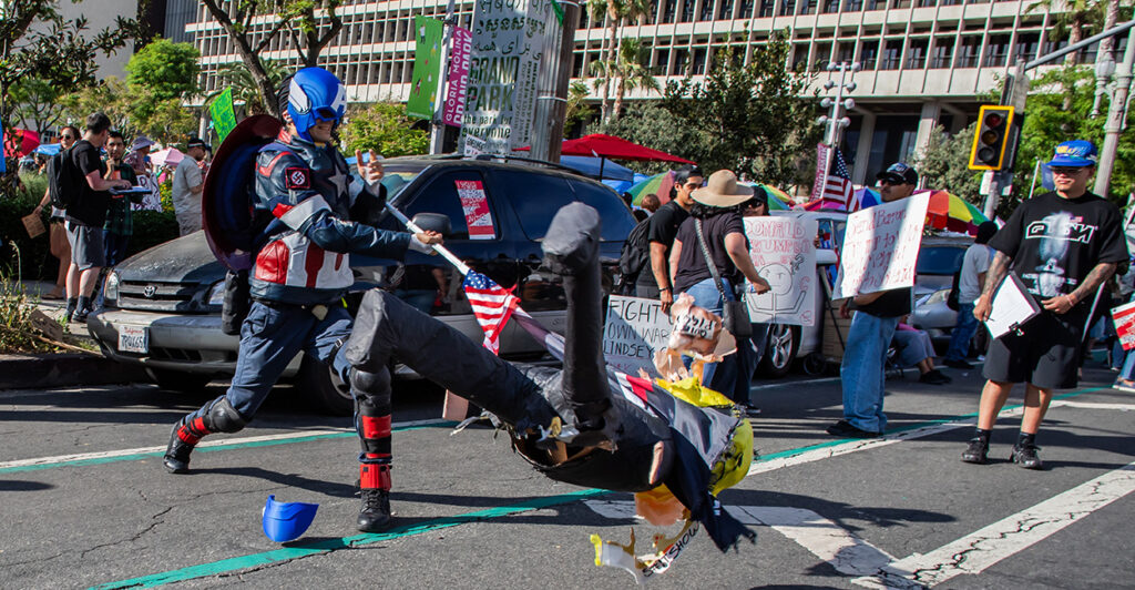 A protester dressed as Captain America impales a human figure with an American flagpole in the middle of the street during a protest.