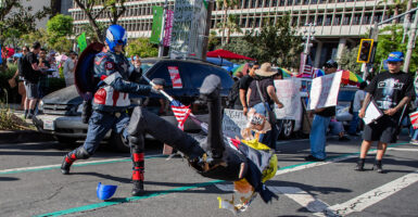 A protester dressed as Captain America impales a human figure with an American flagpole in the middle of the street during a protest.