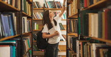 Female student reaching up for a book in a school library stack.