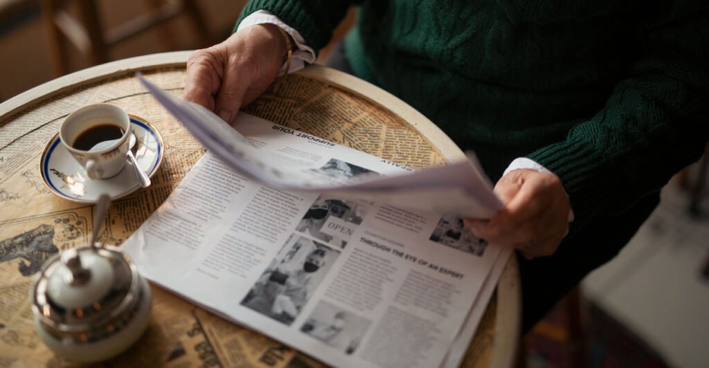 Unseen older person reading a daily newspaper while sitting at round table with cup of coffee
