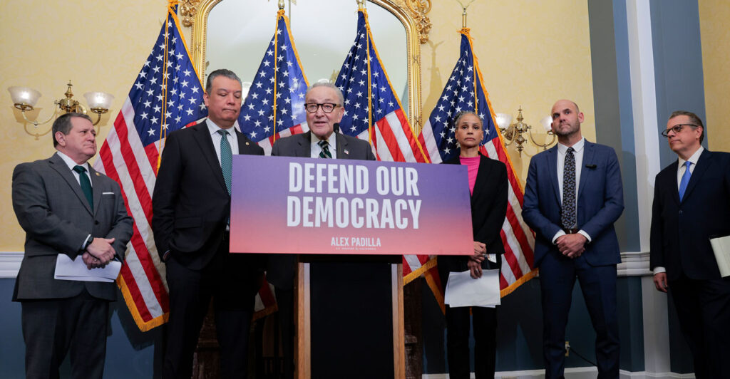 Chuck Schumer and others stand behind a podium which reads "Defend Our Democracy"