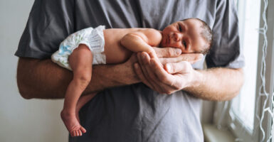 Male in gray T-shirt holds a newborn, whose legs dangleoff his arm.