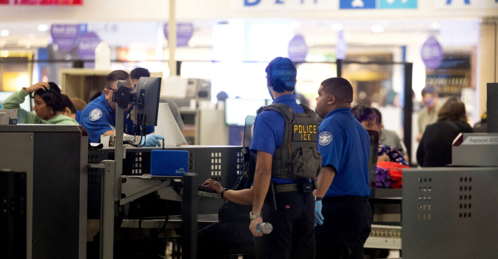 TSA agents assisted by ICE agents screen passengers at an airport.