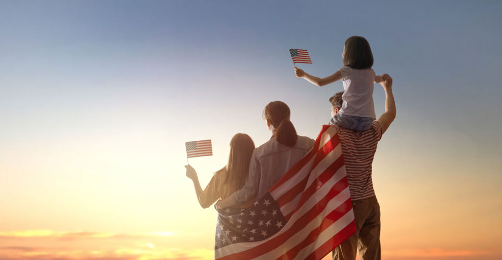 Parents and two daughters, draped in a flag, with the girls waving little flags, look at a beautiful sunset.