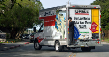 A man in black chats on his cellphone while standing on the back ledge of a Uhaul truck that's pulling out on a suburban street.